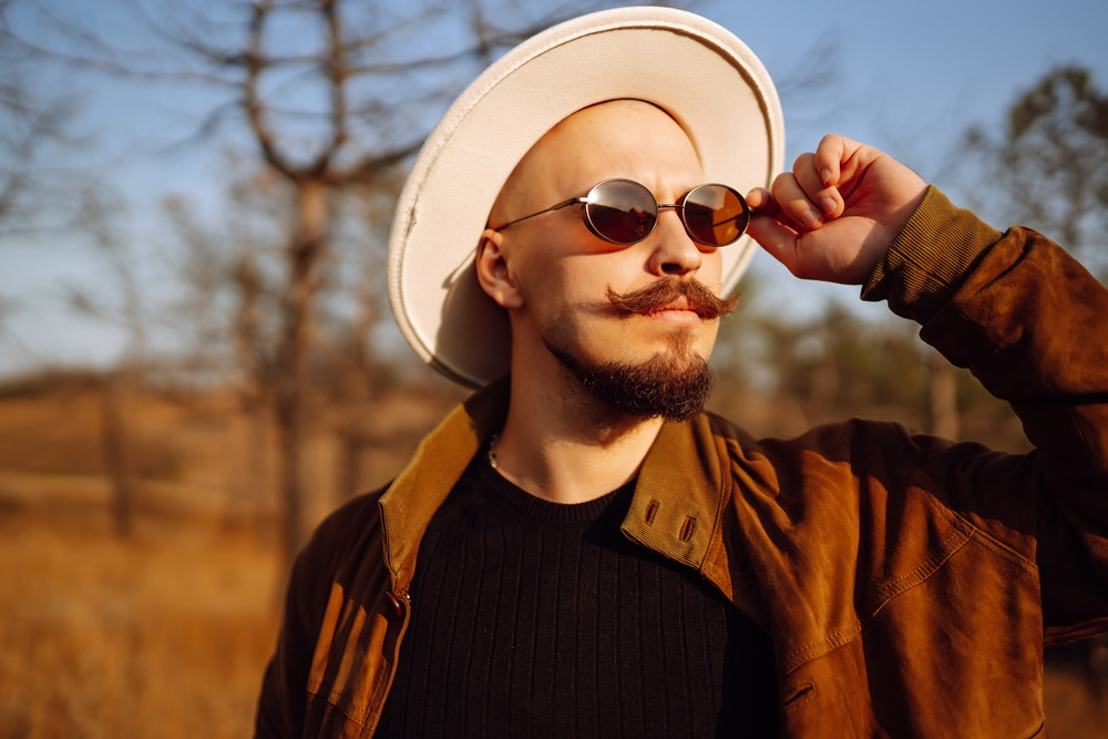 Man with a mustache and beard wearing sunglasses, a white hat, and brown jacket outdoors in sunlight.